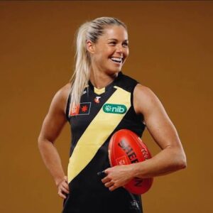 Female athlete with a blonde ponytail, wearing a black and gold diagonal-striped jersey, smiling while holding a red football against a warm brown background.