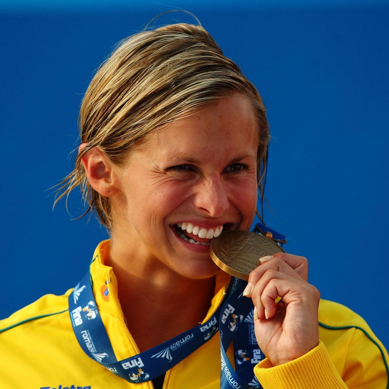 Female athlete in a yellow jacket smiling while biting a bronze medal against a blue backdrop.