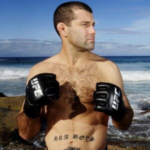 Man shirtless on a rocky beach wearing black UFC gloves, fists up and looking to the right, ocean waves in the background.