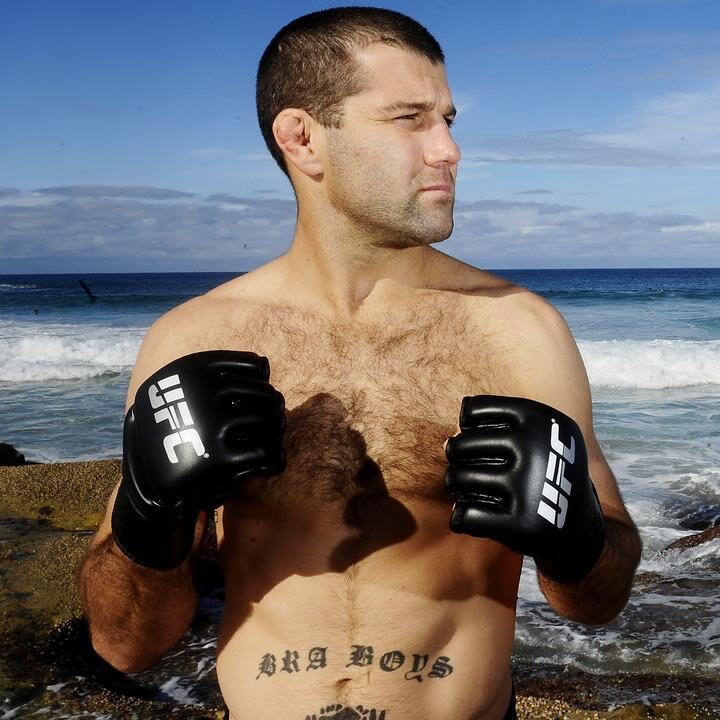Man shirtless on a rocky beach wearing black UFC gloves, fists up and looking to the right, ocean waves in the background.