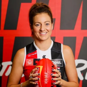 Female athlete in a sleeveless jersey smiles at the camera while holding a red ball in front of a bold red-and-black backdrop, with team logos on her jersey.