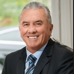 Professional headshot of a smiling older man in a dark suit and blue striped tie, indoors near a window.