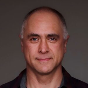 Close-up portrait of a middle-aged man with short gray hair and a neutral expression, looking straight ahead.