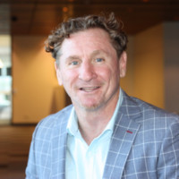 Smiling man in a light blue shirt and blue plaid blazer, photographed in a modern indoor setting (professional headshot).