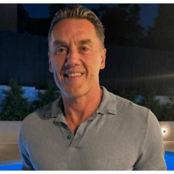 Smiling man in a light blue polo shirt stands beside a lit pool at night with a dark fence in the background.