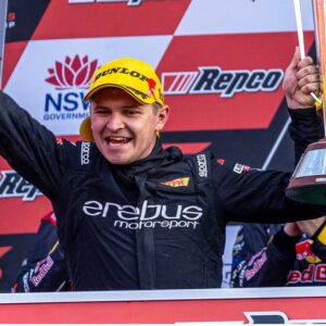 Racing driver in Erebus Motorsport suit on a podium, wearing a yellow Dunlop cap and holding a trophy amid sponsor banners.