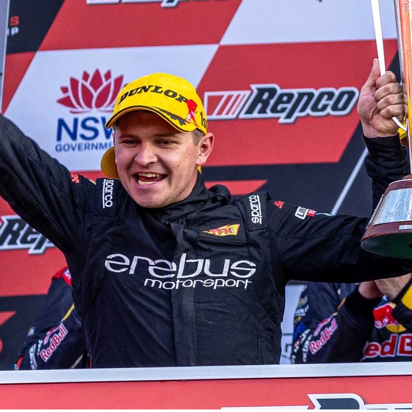 Racing driver in Erebus Motorsport suit on a podium, wearing a yellow Dunlop cap and holding a trophy amid sponsor banners.
