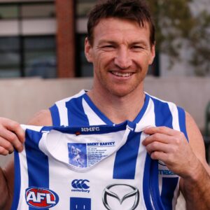 Smiling man in a blue-and-white striped AFL jersey reveals a player card tucked into his shirt.