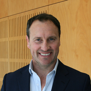 Portrait of a smiling man in a navy suit and light blue shirt, posed against a wood-paneled wall.