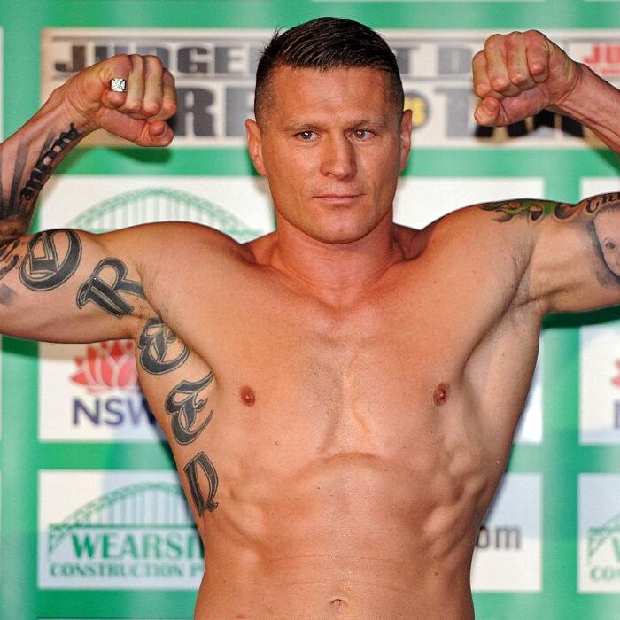 Muscular, shirtless man flexing his biceps on a stage, with tattoos visible on both arms and a green backdrop behind him.