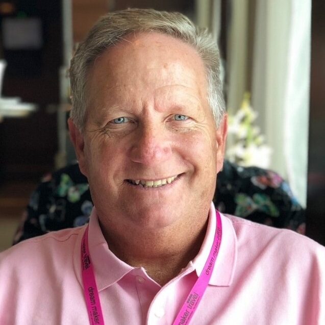 Smiling older man with gray hair in a pink polo shirt and pink lanyard, reading an open book indoors.
