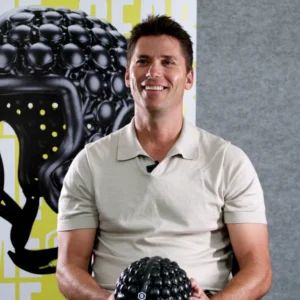 Smiling man in a light polo shirt sits indoors, holding a black textured ball in his lap, with a poster behind him.